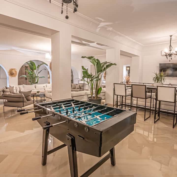 Grand salon clair avec baby-foot, canapés et arches blanches.